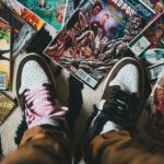 Guardiões da galáxia. From above crop person wearing brown trousers and sneakers standing on pile of collection of comics magazines with colorful illustrations on cover